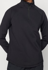 Mann trägt einen schwarzen Pullover mit langen Ärmeln, hohem Kragen und kurzem Reißverschluss vorne, Hände hinter dem Rücken.