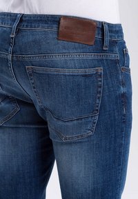Blue denim jeans with a fitted design, featuring rear pockets and a brown leather label at the waistband, showcasing a smooth texture.