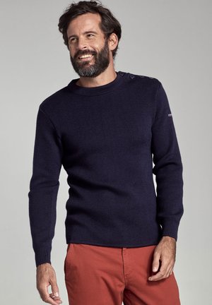Marineblauwe gebreide sweater met een ronde hals en schouderknopen. Heeft ribgebreide manchetten en een ribgebreide hem. Gecombineerd met rode broek.