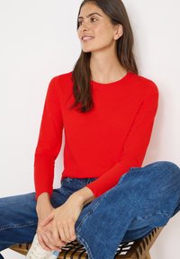 Rotes langärmliges Oberteil in taillierter Passform, aus glattem Stoff. Kombiniert mit blauen weitgeschnittenen Jeans, sitzt auf einem Holzschemel.