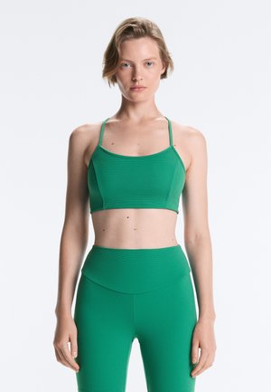 Kobieta z krótkimi blond włosami, ubrana w zielony sportowy stanik i legginsy z wysokim stanem, stojąca na jednolitym jasnym tle.