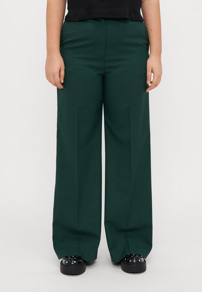 Pantaloni largi verzi cu o textură netedă, având talie înaltă și pliseuri frontale. Stilizați cu pantofi cu platformă negri.