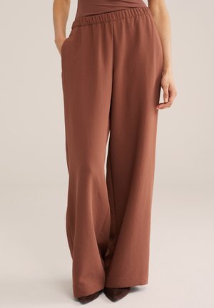 Žena nosí hnedé nohavice s vysokým pásom a širokými nohavicami, s elastickým pásom a bočnými vreckami, doplnené o tmavohnedé špicaté topánky.