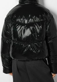 Černá prošívaná bunda s lesklým, lesklým povrchem. Má prošívaný vzor a vysoký límec, který dodává vzhled polstrované, izolované bundy.