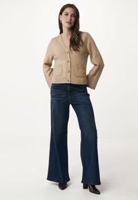 Beige kofta med V-ringning, framknappar och två påsydda fickor; kombinerad med vida mörka jeans och spetsiga klackskor.