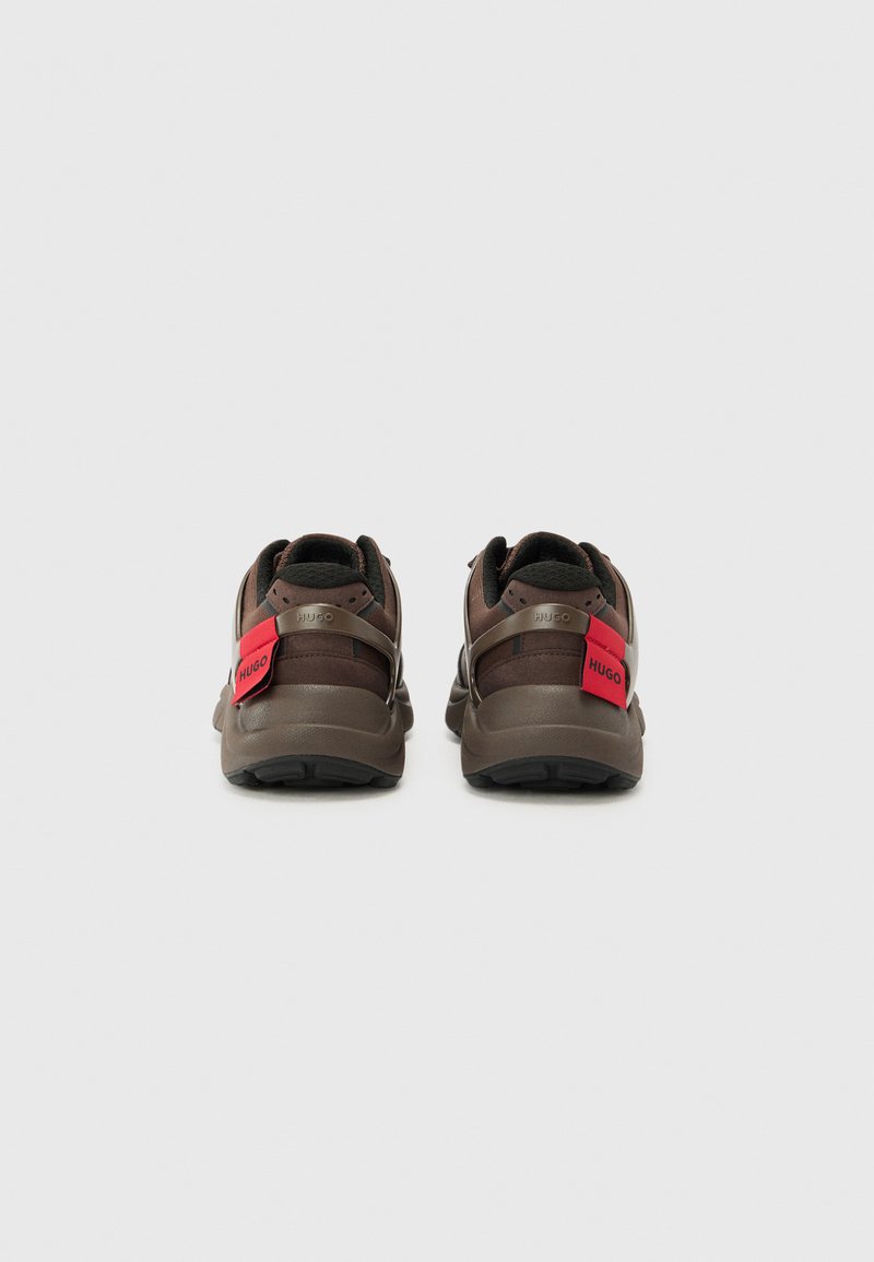 Zapatillas marrones de malla y material sintético, con suela acolchada, correas flexibles y un detalle del logo rojo en el talón.