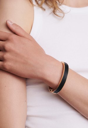 Schwarzes Lederarmband mit goldfarbener Metallverzierung, am Handgelenk getragen, vor einem weißen, gerippten Tanktop-Hintergrund, der glatte Texturen und kontrastierende Farben zeigt.