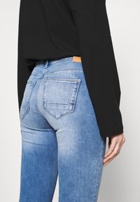 Jeans de mezclilla azul con un lavado desgastado, diseño de cinco bolsillos y una etiqueta de cuero marrón en la parte trasera. Acompañados de una camiseta de manga larga negra.