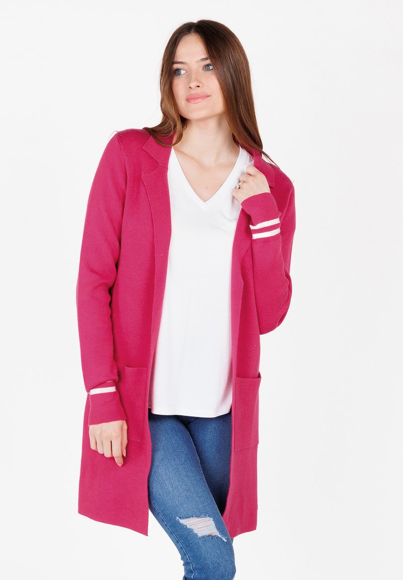 Cardigan lungo fucsia con due tasche frontali, apertura anteriore e polsini a righe bianche, indossato sopra una maglietta bianca e jeans blu. Texture liscia.