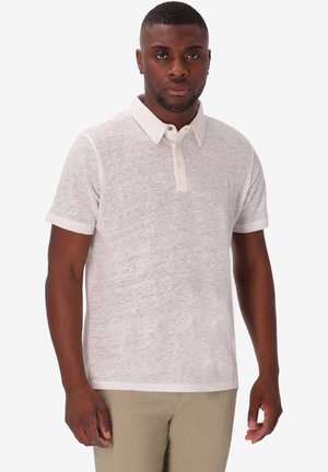 Uomo con capelli corti che indossa una polo bianca a maniche corte e pantaloni beige, in piedi davanti a uno sfondo chiaro uniforme.