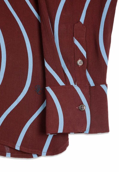Poignet de chemise marron à manches longues avec des rayures courbes bleues, deux boutons et un petit logo brodé "BC" près du poignet.