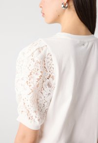 Tricou alb cu mâneci bufante din dantelă florală, având un corp din bumbac neted. Detalii subtile la spate, cu o mică aplicație.