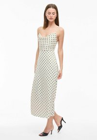 Robe midi couleur crème avec des pois noirs, fines bretelles spaghetti, corsage ajusté et silhouette droite ; le tissu semble lisse et léger.