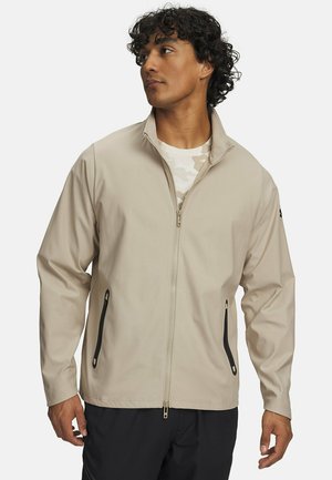 Mann trägt eine beige Jacke mit Reißverschluss und schwarzen Reißverschlusstaschen sowie ein helles gemustertes Hemd darunter und schaut seitlich.
