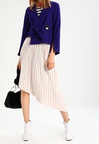 Fialová krátká bunda s detailem na zavazování, krémová plisovaná asymetrická sukně a bílé kotníkové boty. Černá kabelka visí na paži.