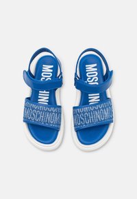 Blå sandaler med blank yta, med ett mönstrat rem med "MOSCHINO"-text och en vit, vadderad sula. Justerbar sidrem.