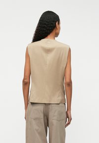 Bez piedurknēm beige tops, izgatavots no gludas auduma; strukturēta dizaina ar augstu kakla izgriezumu un smalku aizmugures šuvju detaļu, apvienots ar beige biksēm.