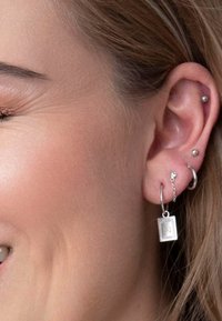 Zilveren oorbellen met een hangende rechthoekige bedel met een gegraveerd patroon. Meerdere oorpiercingen zichtbaar. Natuurlijke haarkleur.