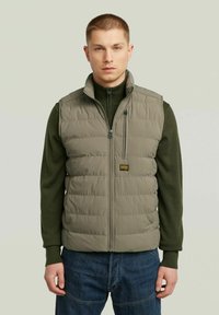 Gilet leggero e imbottito in un verde oliva soft, con zip frontale, colletto alto e patch con logo. Indossato sopra un maglione di maglia scura.