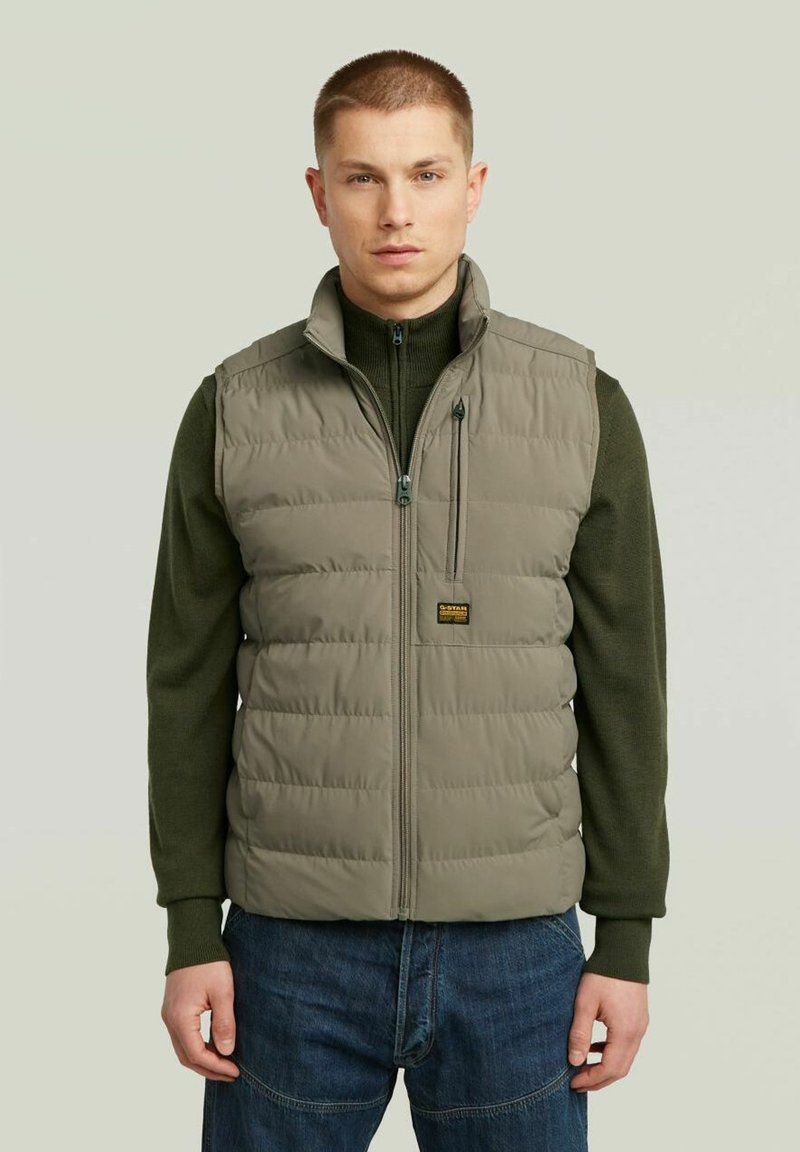 Gilet leggero e imbottito in un verde oliva soft, con zip frontale, colletto alto e patch con logo. Indossato sopra un maglione di maglia scura.