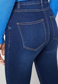 Dunkelblaue Jeans aus Denim mit hoher Taille, ausgestattet mit zwei Gesäßtaschen und kontrastierendem orangefarbenen Nähten. Glatte Textur mit leichten Verwaschungen.
