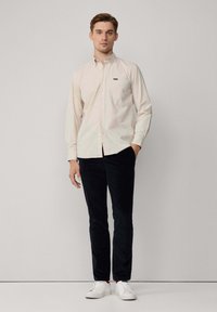 Lichtbeige overhemd met knopen en een borstzak, gecombineerd met donkere corduroy broek en witte sneakers. Eenvoudig, strak ontwerp en pasvorm.