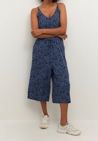 Une femme se tient les bras croisés, vêtue d'une combinaison sans manches à imprimé léopard bleu et de baskets blanches épaisses, devant un mur uni.