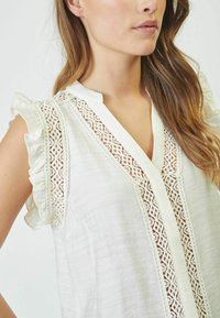 Blusa bianca con dettagli in pizzo lungo il collo e le spalle, con maniche corte arricciate e una vestibilità rilassata.