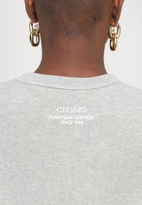 Pilkas megztinis su apvalia iškirpte, nugaroje su baltu tekstu, kuris skamba „UŽDARYTA EUROPOS PAVELDAS NUO 1978 M.“ Minkšta tekstūra.