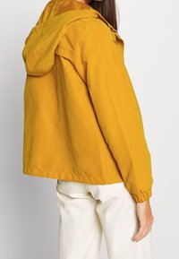 Żółta kurtka z kapturem o luźnym kroju, elastycznych mankietach i kapturze ze sznurkiem. Lekka tkanina, widoczne detale przeszyć, minimalistyczny design.
