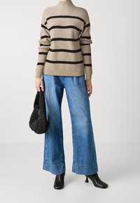 Beżowy sweter w paski z czarnymi poziomymi prążkami, zestawiony z szerokimi niebieskimi dżinsami z dużym stanem. Trzyma czarną plecioną torbę.