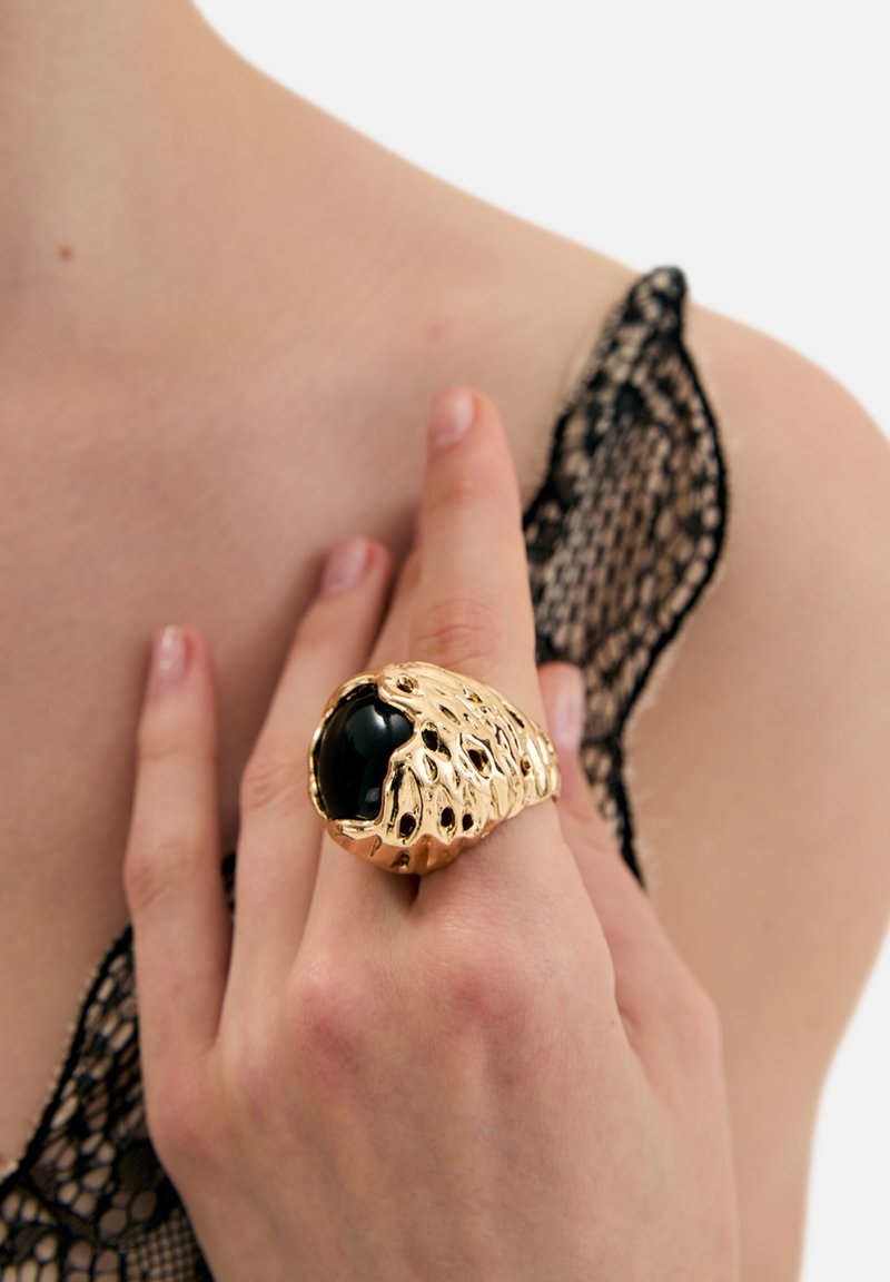 Anillo abstracto de oro con superficie texturizada y múltiples agujeros, engastado con una gran piedra negra. Mano modelando el anillo sobre piel desnuda.
