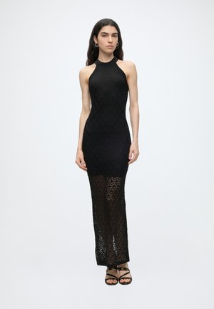 Femme aux longs cheveux foncés portant une robe maxi noire en crochet sans manches et des sandales noires à lanières, debout devant un fond clair uni.