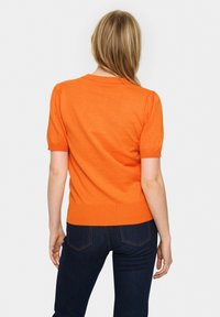Pulover tricotat cu mânecă scurtă, de culoare portocalie, cu tiv ondulat și detalii cu mâneci bufantă, asortat cu blugi din denim închisi. Design simplu și texturat al materialului.