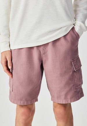 Mann trägt hellviolette Cargo-Shorts mit Seitentaschen und ein weißes, strukturiertes Langarmshirt, die Hände ruhen in der Nähe der Taschen.