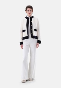 Weiße Tweedjacke mit schwarzen Akzenten, verziert mit goldenen Knöpfen, fransigen Kanten und zwei vorderen Taschen. Kombiniert mit weißen Glockenhosen.