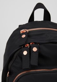 Sac à dos noir avec fermetures éclair en or rose et tirettes en tissu noir ornées de boutons métalliques ronds, montrant une poignée supérieure et deux compartiments avant.