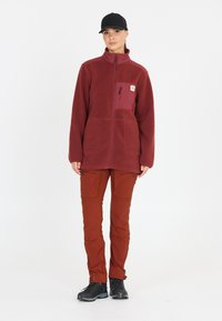 Burgundy-värinen fleece-takki, jossa on rintatasku, vetoketjukiinnitys, kaksi etutaskua, yhdistetty ruosteenvärisiin housuihin ja mustiin vaelluskenkiin.