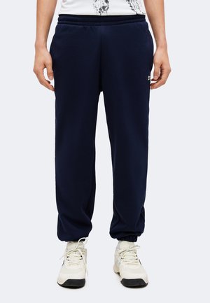 Persoană îmbrăcată în pantaloni sport bleumarin cu manșete elastice și adidași albi, stând pe un fundal simplu.