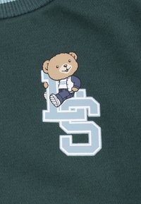 Dunkelgrüner Stoff mit einem cartoonartigen Bärendesign und hellblauem "LS"-Text. Der Bär trägt eine Jacke und sitzt auf dem Buchstaben "L".