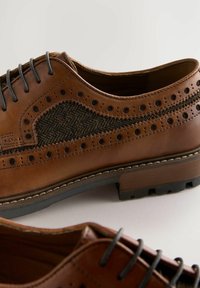 Chaussures en cuir marron avec un panneau en tissu noir texturé, des détails perforés, des lacets plats et une semelle en caoutchouc robuste avec un motif de traction visible.