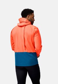 Veste à coloris contrastés présentant une section supérieure orange et une section inférieure bleue. Le matériau semble lisse et dispose d'une capuche ainsi que de poignets élastiques.