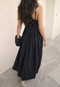 Svart maxiklänning med veckad överdel och flödande kjol. Kompletterad med en texturerad svart vävd handväska. Bakgrunden har ljusa väggar.