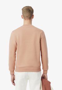 Sudadera de melocotón claro con cuello redondo y puños acanalados, combinada con pantalones blancos, sostenida con una chaqueta de exterior en un brazo.