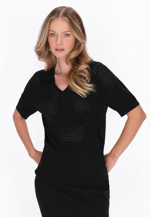 Kvinde med langt, bølget blondt hår iført en sort, kortærmet, perforeret V-hals top, stående med hænderne på hoften mod en hvid baggrund.