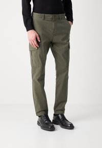 Pantalones cargo verde oliva con bolsillos laterales, un ajuste regular y cierre de botón. La tela tiene una textura suave y resistente. Unos zapatos negros completan el look.