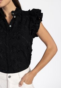 Blouse noire ornée de broderies florales, manches courtes à volants et col avec fermetures par boutons. Le tissu présente une finition texturée.