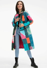Barevný patchworkový kabát s různými vzory v odstínech modrozelené, růžové a oranžové; dlouhý design s knoflíky a podšívkou v červené barvě.