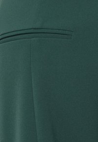 Tecido verde com uma textura suave. Apresenta um detalhe de costura horizontal e um design limpo e minimalista. Nenhum padrão ou peças de hardware visíveis.