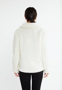 Hvid, tekstureret fleecejakke med høj krave og lange ærmer. Har en afslappet pasform og buet kant, parret med sorte leggings.